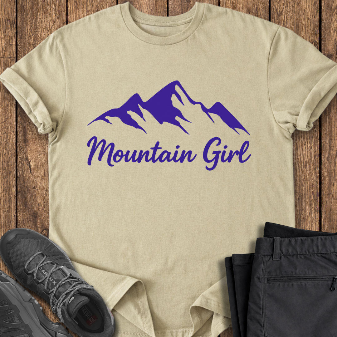 Beige t-shirt with purple mountain graphic and 'Mountain Girl' text on a wooden surface with black shoes and pants.