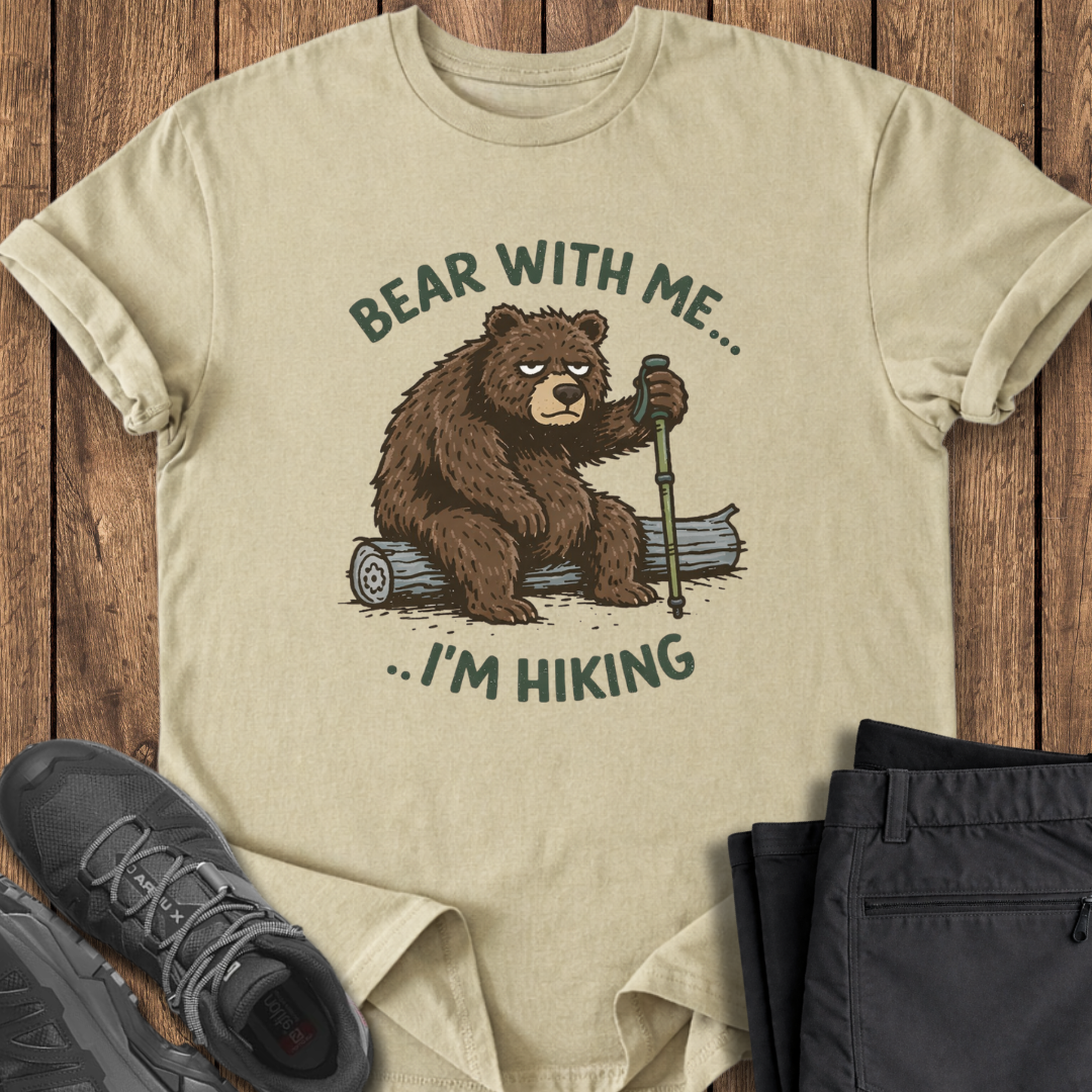 Beige t-shirt with bear graphic and text 'Bear with me... I'm hiking' on a wooden surface with shoes and pants. slow hiking funny t shirt grumpy bear hiker