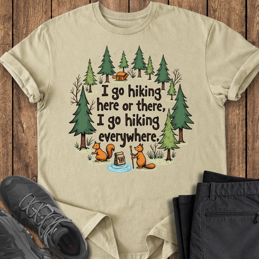 Beige t-shirt with hiking-themed design featuring trees, a cabin, and squirrels on a wooden surface.