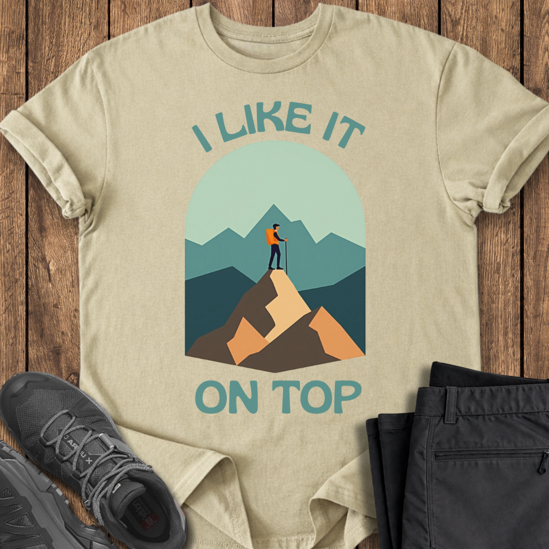 Beige t-shirt with mountain graphic and text 'I Like It On Top' on a wooden surface with shoes and pants.