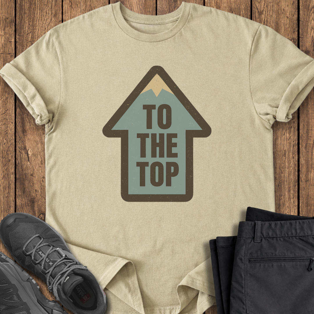 Beige t-shirt with 'To The Top' graphic on a wooden surface with shoes and pants.