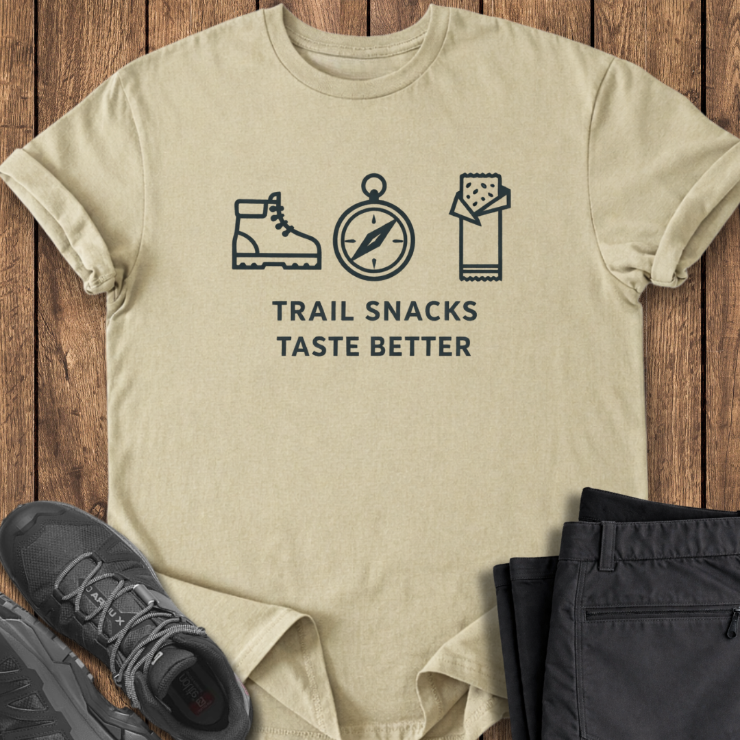 Beige t-shirt with outdoor-themed graphics and text on a wooden surface with shoes and pants. trail snacks taste better