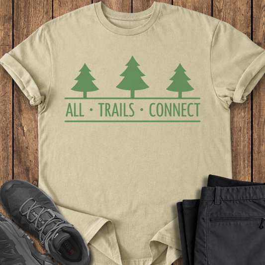 Beige t-shirt with green tree design and 'All Trails Connect' text on a wooden surface with black pants and shoes.