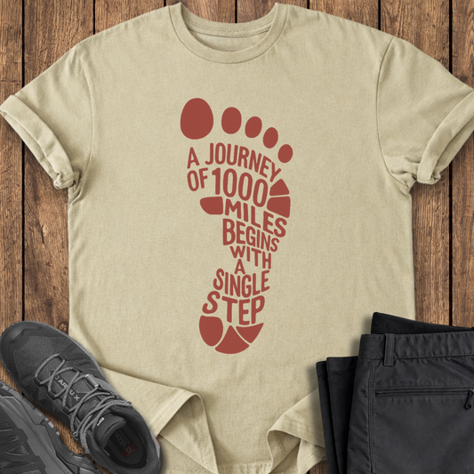 Beige t-shirt with red footprint design and text on a wooden surface, accompanied by black sneakers and pants.