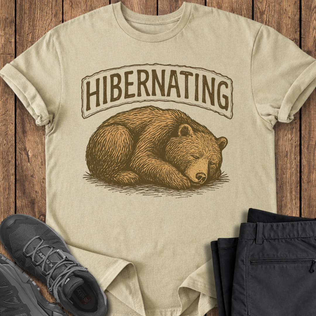 Beige t-shirt with bear graphic and 'Hibernating' text on a wooden surface with shoes and pants.