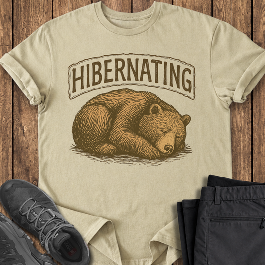 Beige t-shirt with bear graphic and 'Hibernating' text on a wooden surface with shoes and pants.