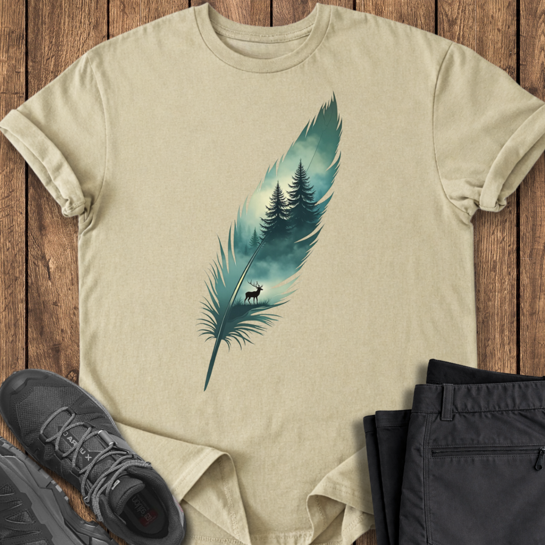 Beige t-shirt with a feather design featuring trees and a deer, placed on a wooden surface with shoes and pants.