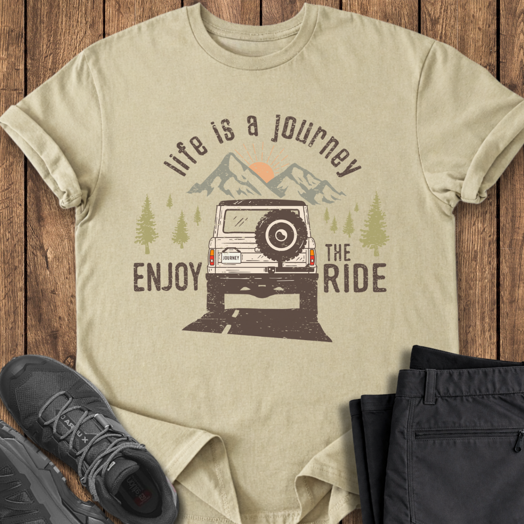 Beige t-shirt with graphic design and text 'Life is a journey, enjoy the ride' on a wooden surface with shoes and pants.