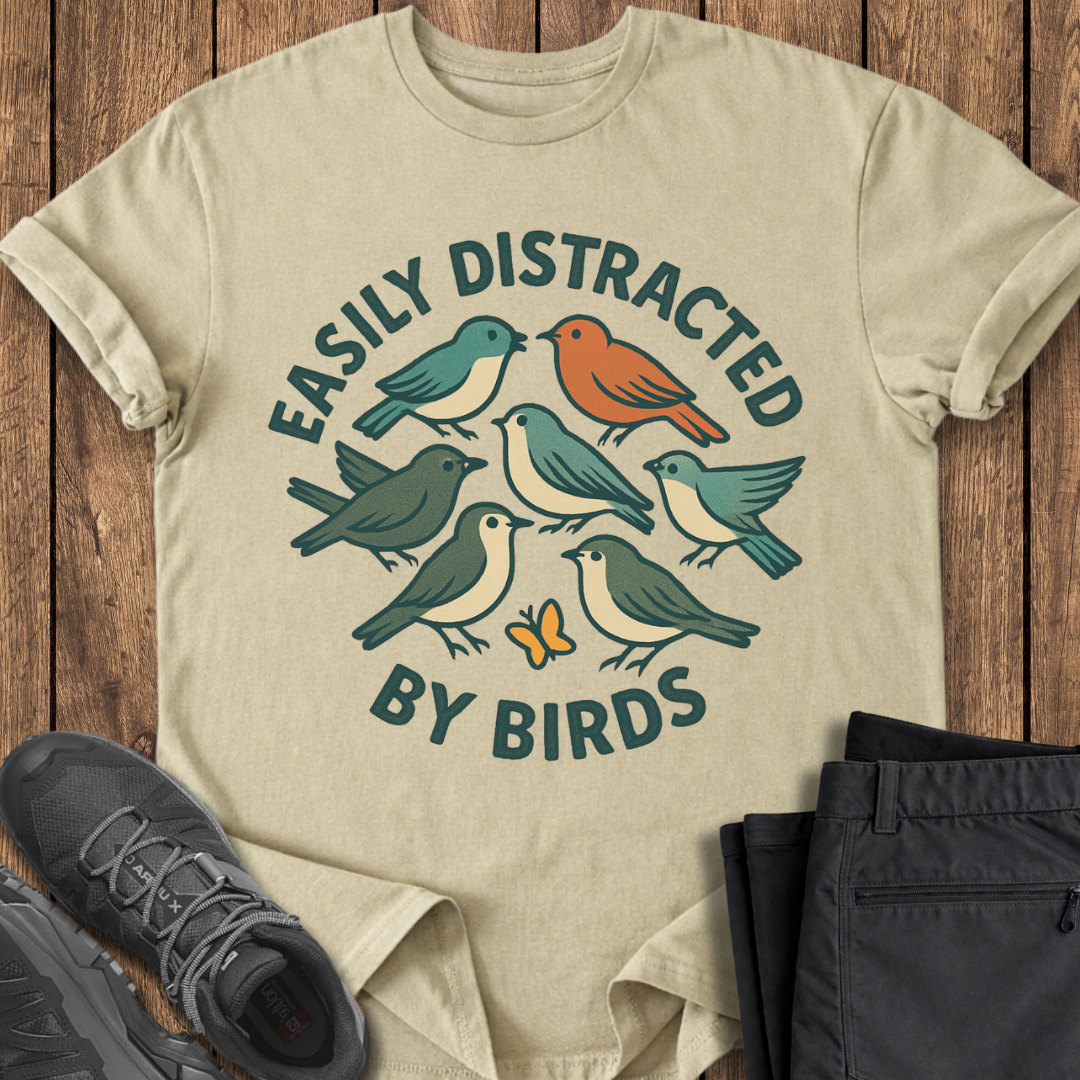 Beige t-shirt with bird design and text on a wooden surface with shoes and pants. easily distracted by birds birding t shirt