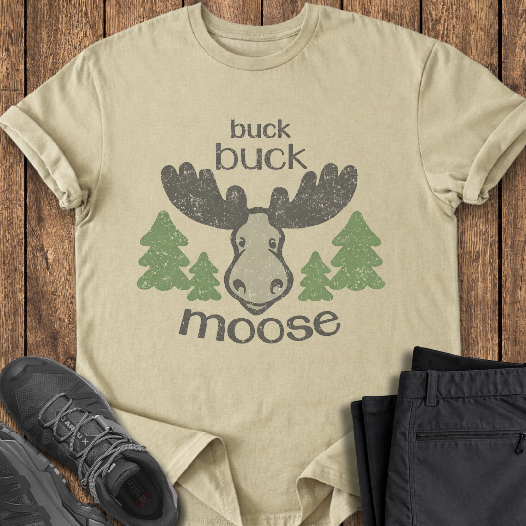 Beige t-shirt with moose graphic and text on a wooden surface with shoes and pants. funny moose t shirt