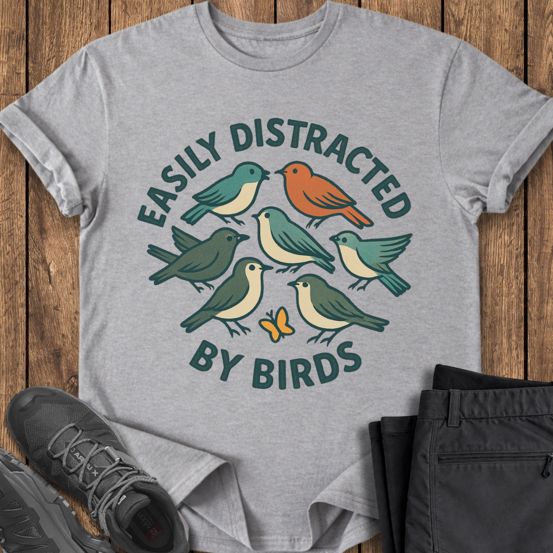 Gray t-shirt with bird design and text on a wooden surface with shoes and pants. easily distracted by birds birding t shirt