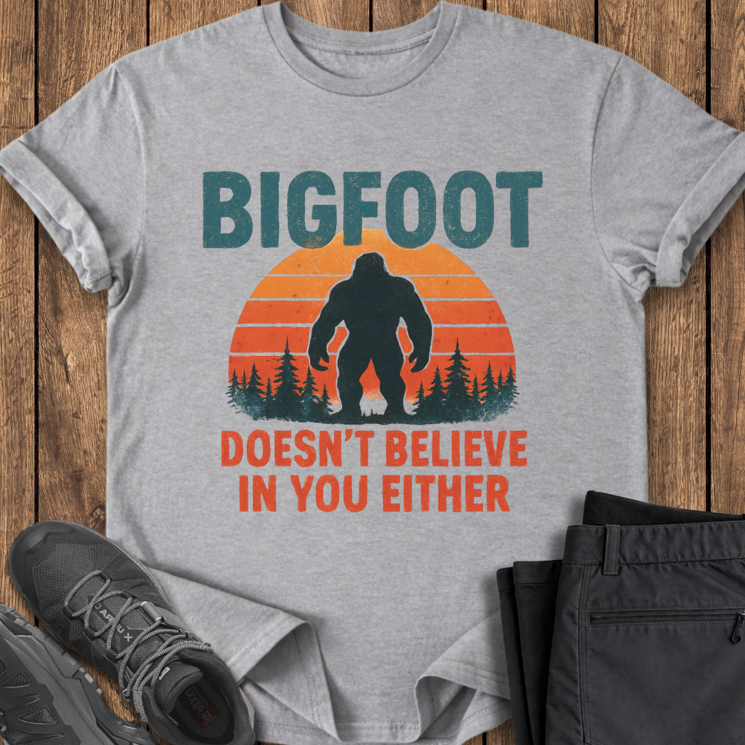 Gray t-shirt with Bigfoot graphic and text on a wooden surface with shoes and pants. funny bigfoot t shirt