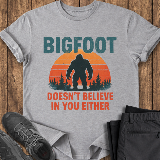 Gray t-shirt with Bigfoot graphic and text on a wooden surface with shoes and pants. funny bigfoot t shirt