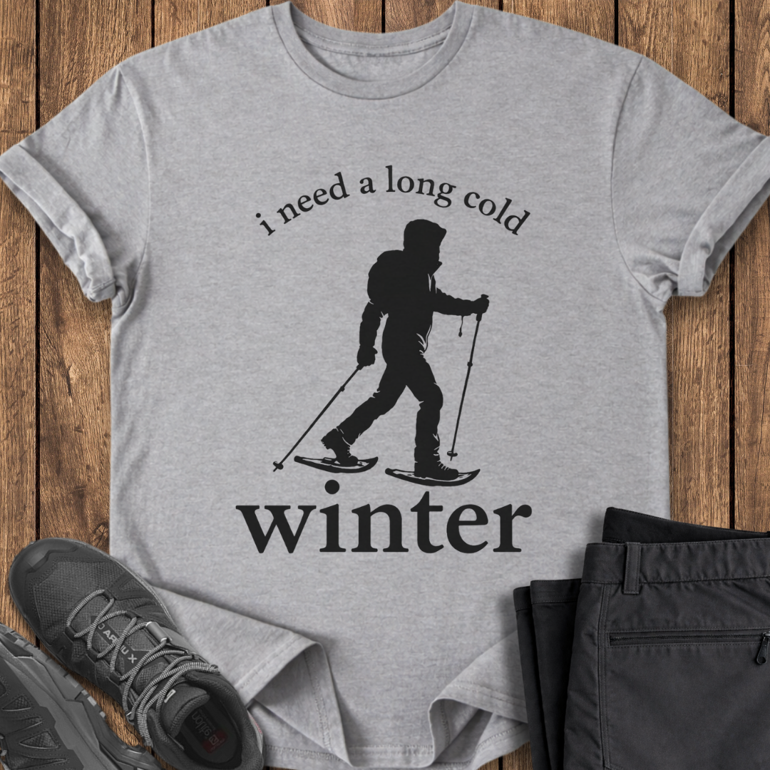 Gray t-shirt with skiing graphic and text 'i need a long cold winter' on a wooden surface with shoes and pants. winter snowshoe  t shirt