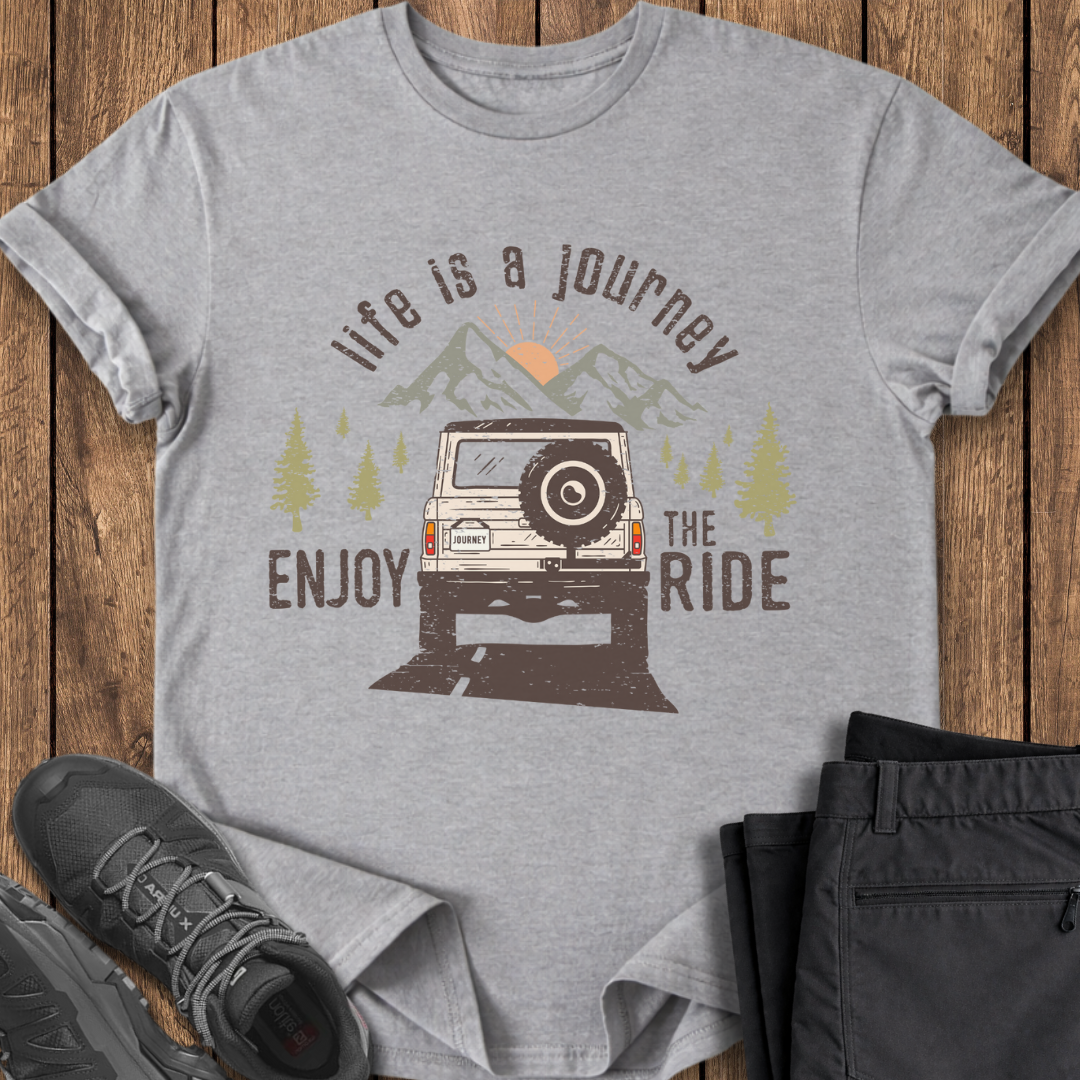 Gray t-shirt with graphic design of a jeep and text 'Life is a journey, enjoy the ride' on a wooden surface.