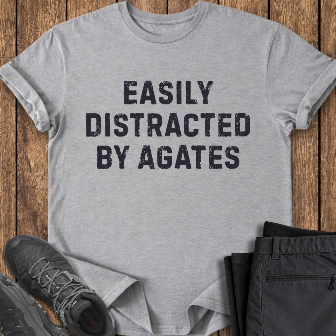 Gray t-shirt with 'Easily Distracted by Agates' text on a wooden surface with shoes and pants.