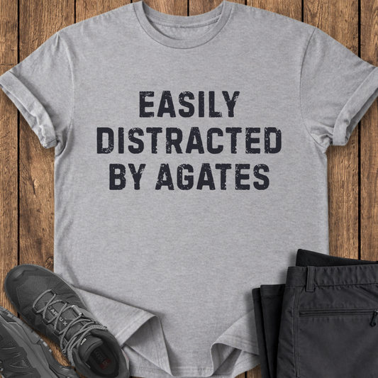Gray t-shirt with 'Easily Distracted by Agates' text on a wooden surface with shoes and pants.