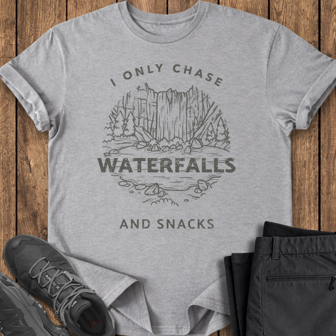 Gray t-shirt with a graphic and text 'I only chase waterfalls and snacks' on a wooden surface with shoes and pants. waterfalls chaser