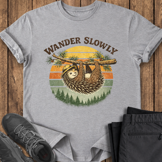 Gray t-shirt with a sloth graphic and 'Wander Slowly' text on a wooden surface with shoes and pants. porcupine hiking tee