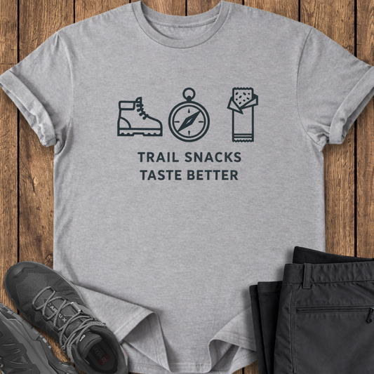 Gray t-shirt with hiking-themed design and text on a wooden surface with shoes and pants. trail snacks taste better