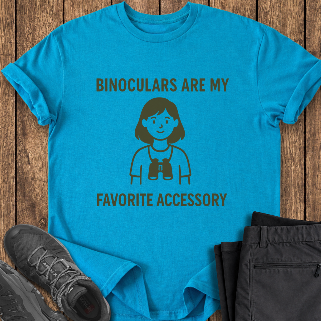 Blue t-shirt with binoculars graphic and text on a wooden surface with shoes and pants.